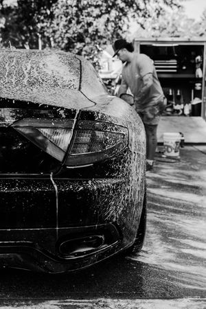 Black and white photo of a soapy sports car being hand washed outdoors by a person near a garage.