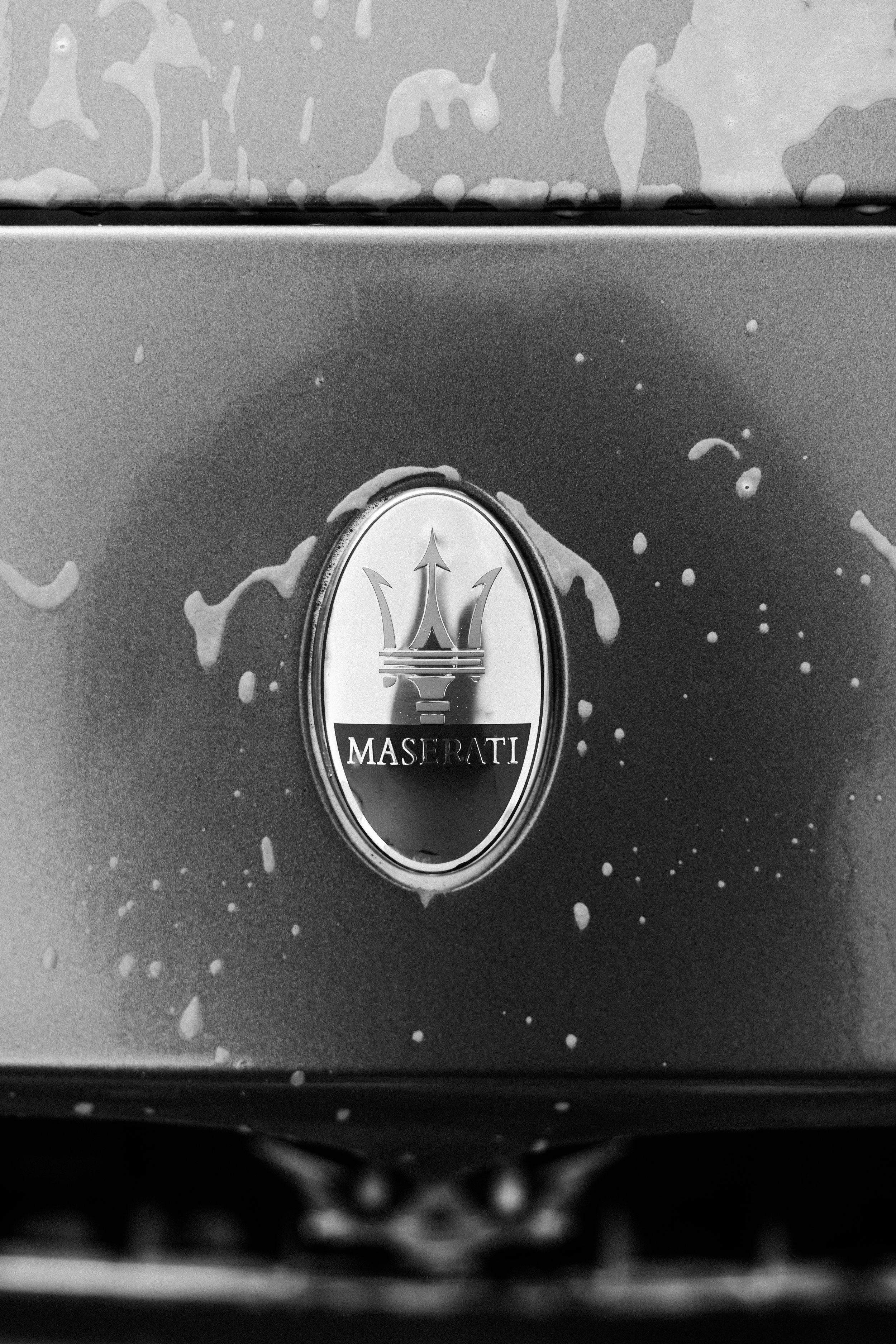 Black and white close-up of a Maserati emblem with trident logo on a wet car hood with water droplets.