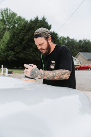 Tattooed man in black t-shirt and cap using an orbital polisher to buff a white car hood outdoors.