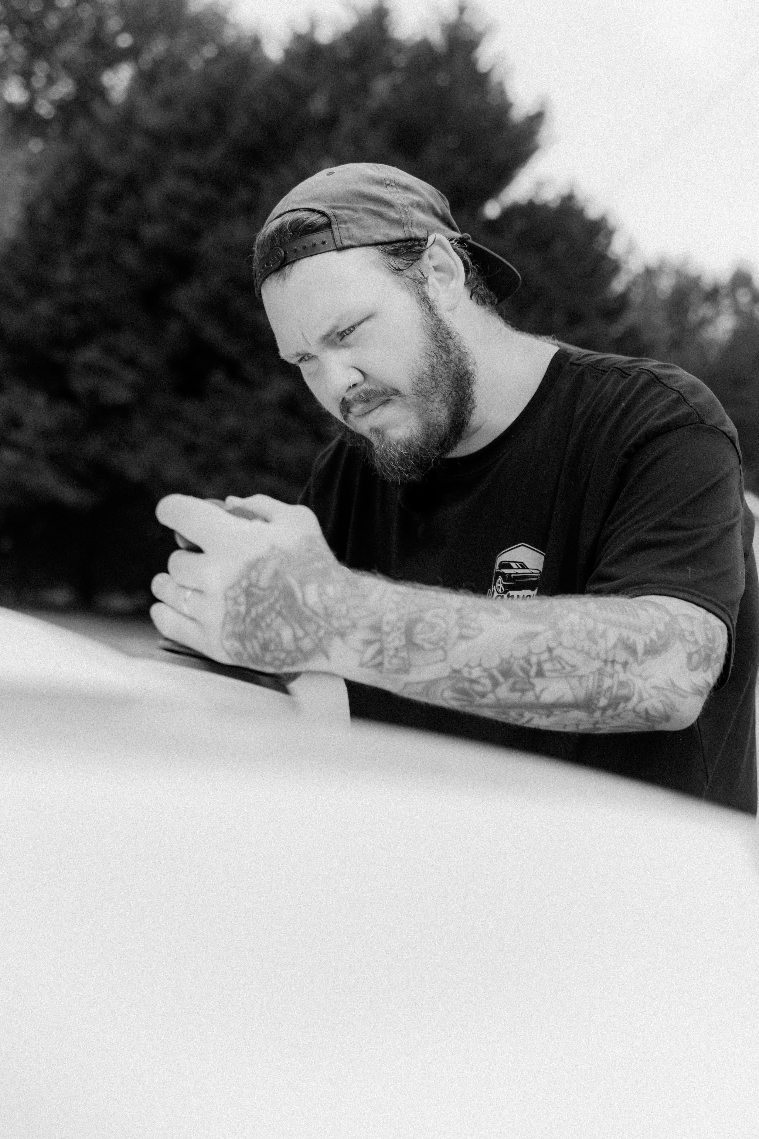 Black and white photo of a tattooed bearded man in a snapback cap leaning on a white car outdoors.