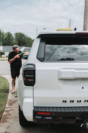 Man pressure washing white Toyota 4Runner SUV on residential driveway.