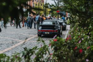Black Porsche 911 parked on cobblestone street with pedestrians and colorful flowers in foreground.
