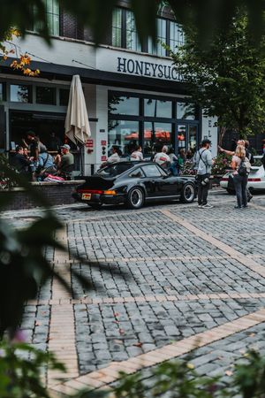 Black Porsche 911 parked on cobblestone street outside Honeysuckle cafe with people dining outdoors.