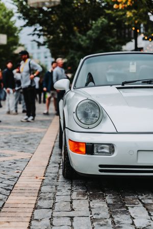 White vintage Porsche 911 parked on cobblestone street with pedestrians in background.