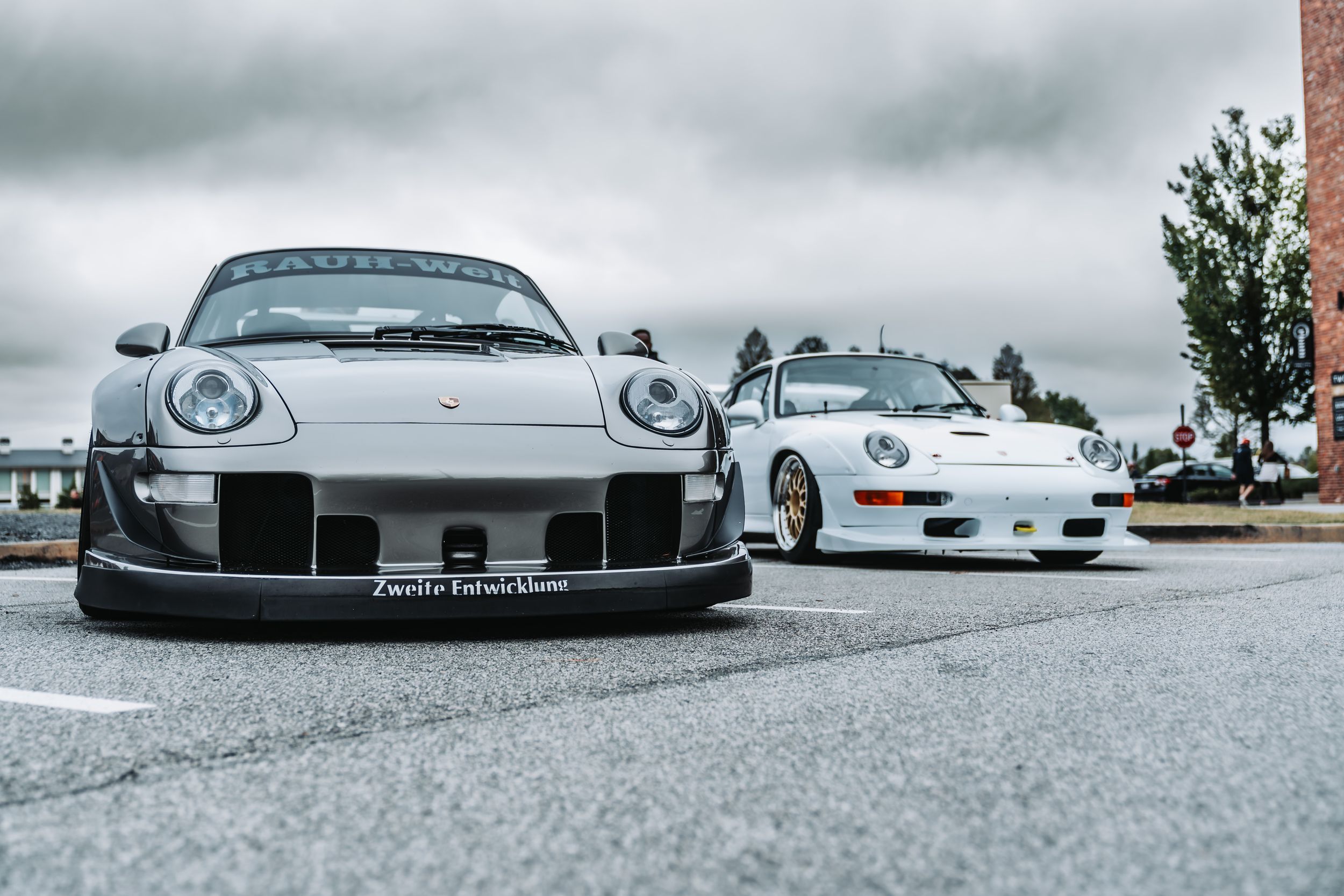Two modified Porsche 911 RWB wide-body kit cars parked on pavement under cloudy skies at a car meet.