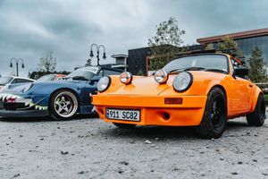 Vintage orange Porsche 911 and blue race car parked at an outdoor car meet on a cloudy day.