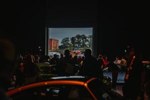 Dark drive-in theater with silhouetted crowd watching a projected outdoor scene with a red car on a large screen.