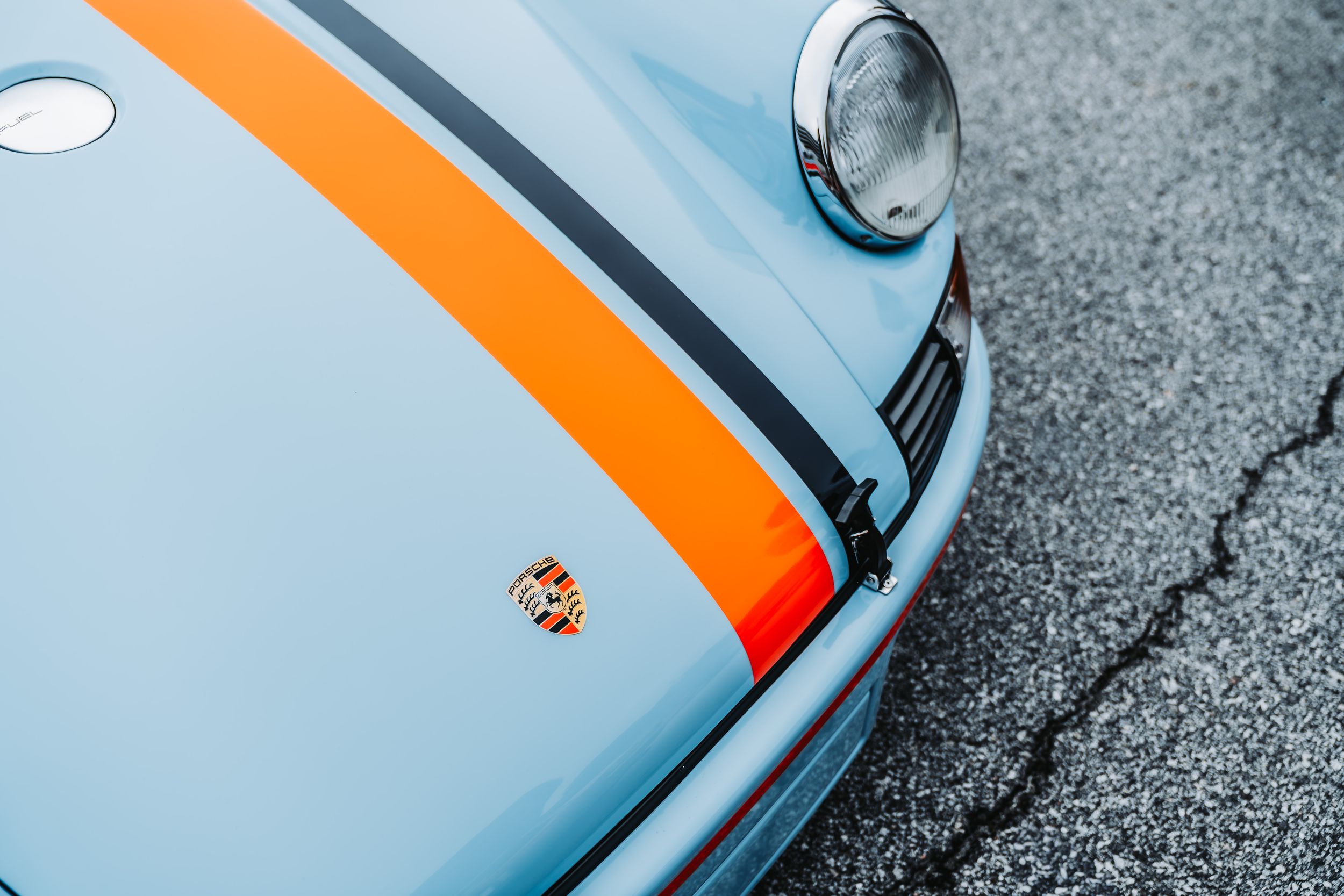 Close-up of a classic Porsche hood featuring Gulf livery blue paint with orange and black racing stripes and badge.