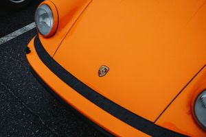 Close-up of a vibrant orange Porsche sports car hood featuring the iconic Porsche crest emblem on asphalt.