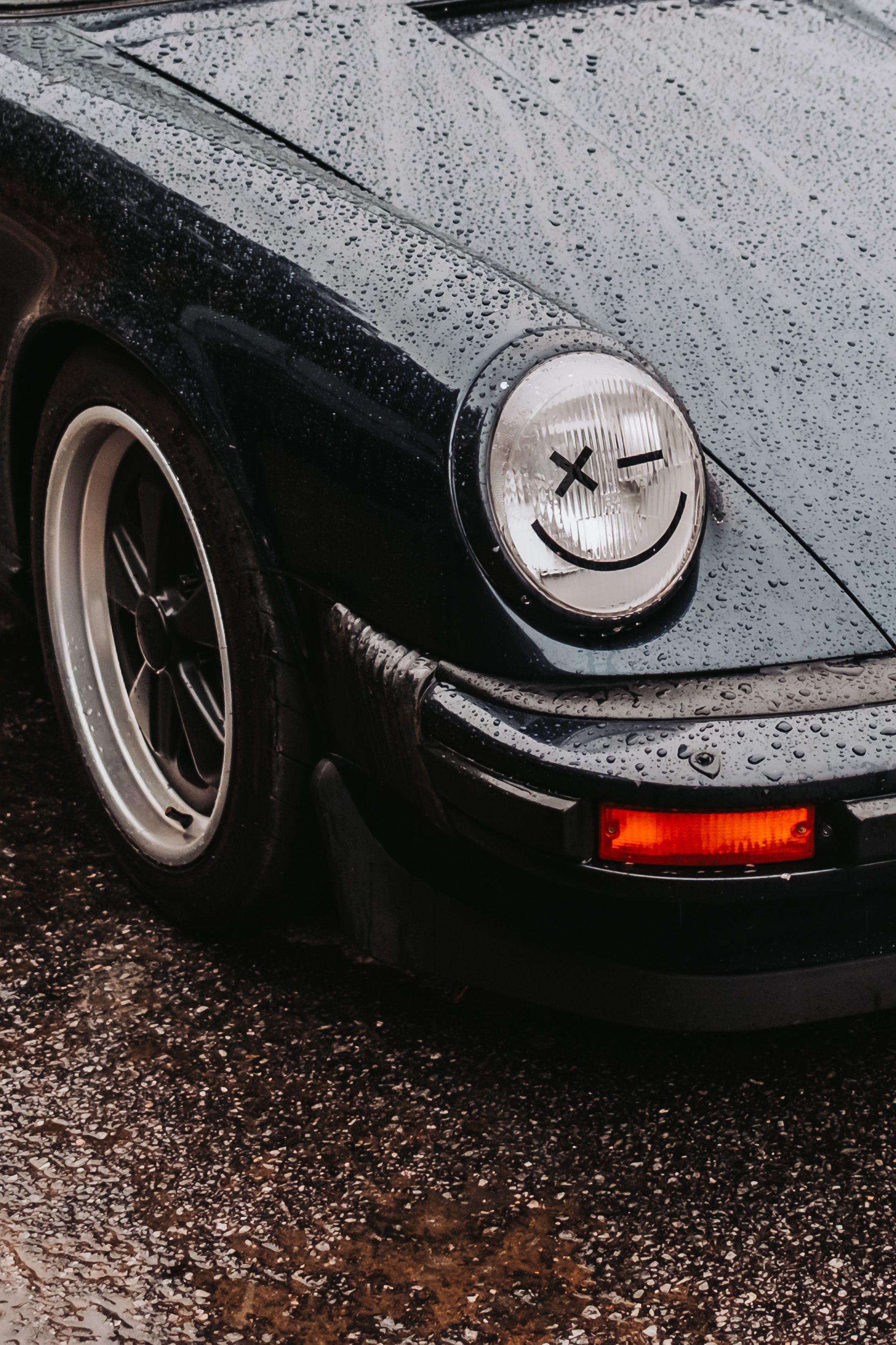A rain-covered black Porsche 911 with a smiley face design on its headlight, parked on wet pavement.