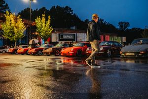 Man walking in rainy parking lot beside a row of classic Porsche sports cars at dusk near a shopping center.