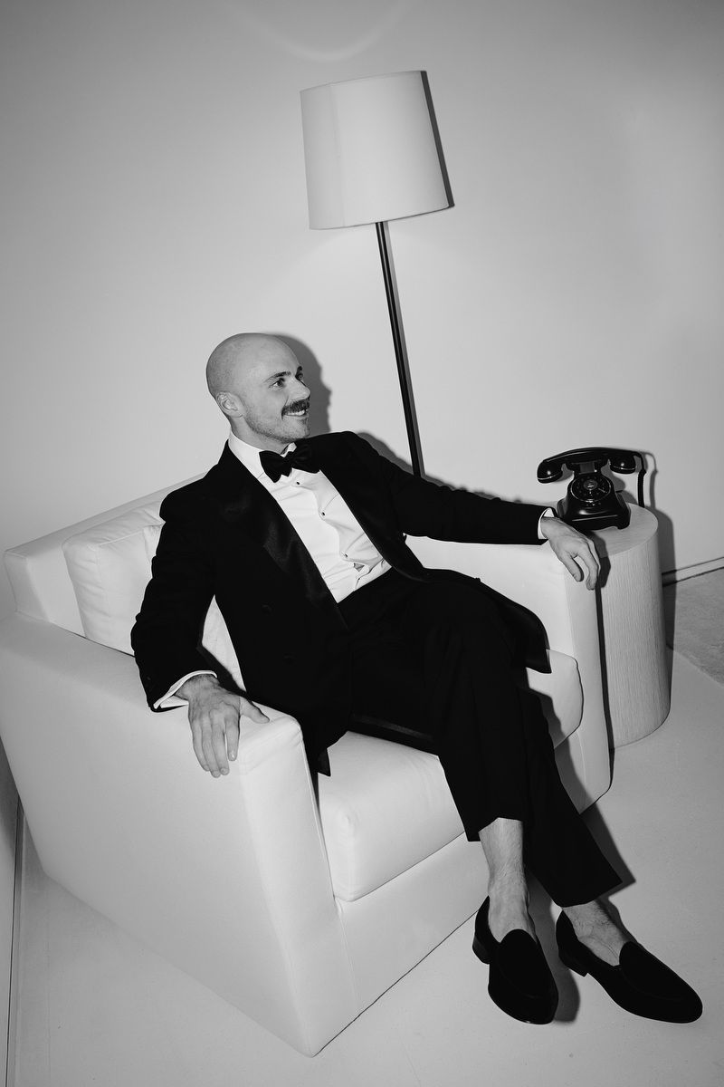 Bald man in tuxedo with bow tie sits in white armchair holding vintage rotary phone beside floor lamp.