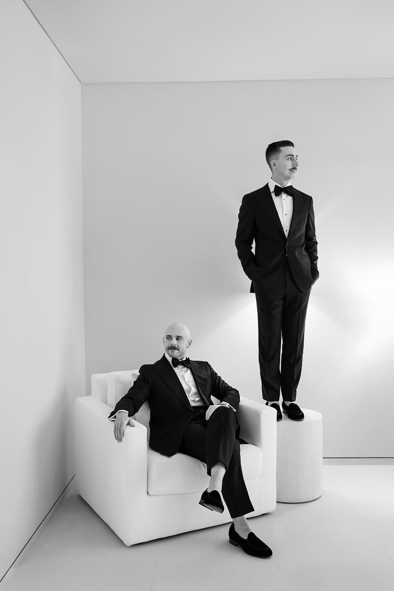 Two men in black tuxedos pose in a minimalist white room, one seated in a chair and one standing on a pedestal.