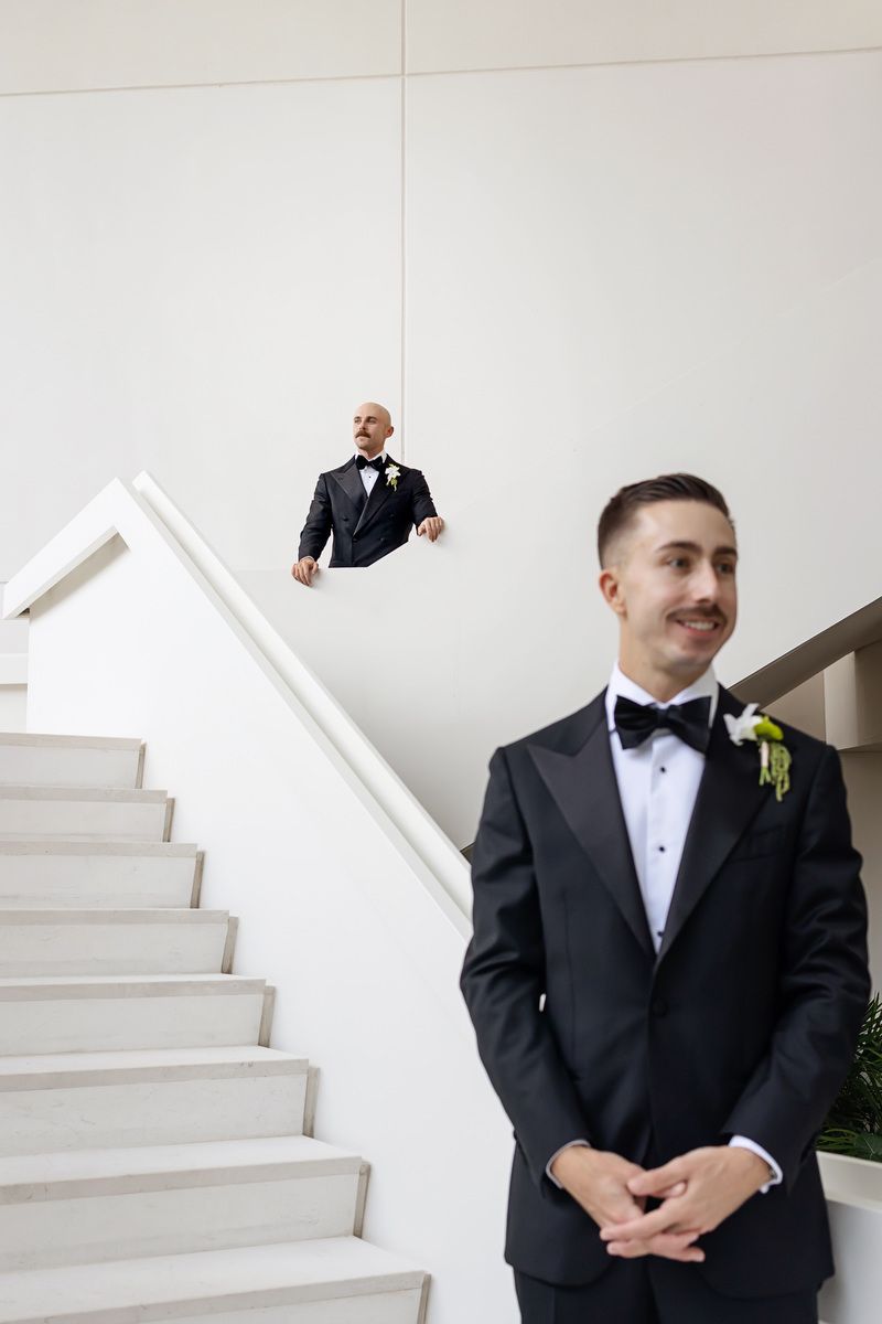 Two grooms in black tuxedos with boutonnieres pose on a staircase in a modern white venue on their wedding day.