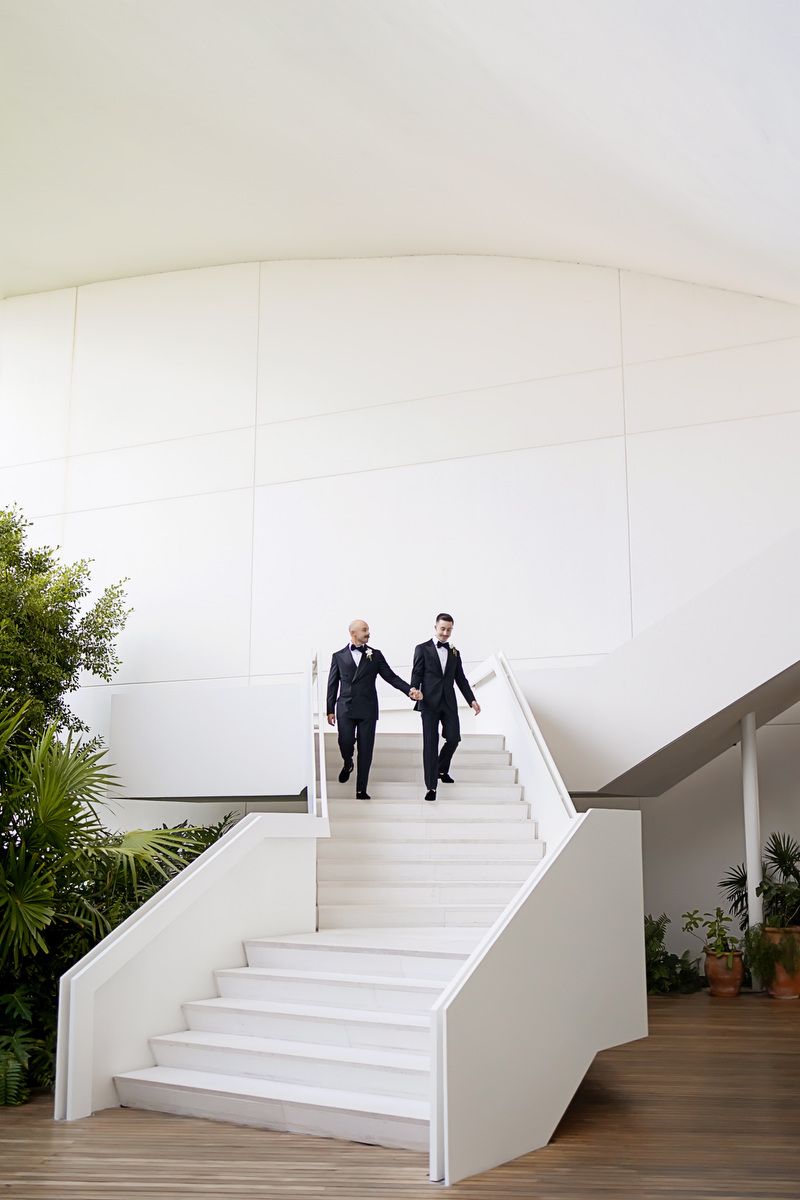 Two grooms in black tuxedos hold hands while descending white stairs at a modern wedding venue.