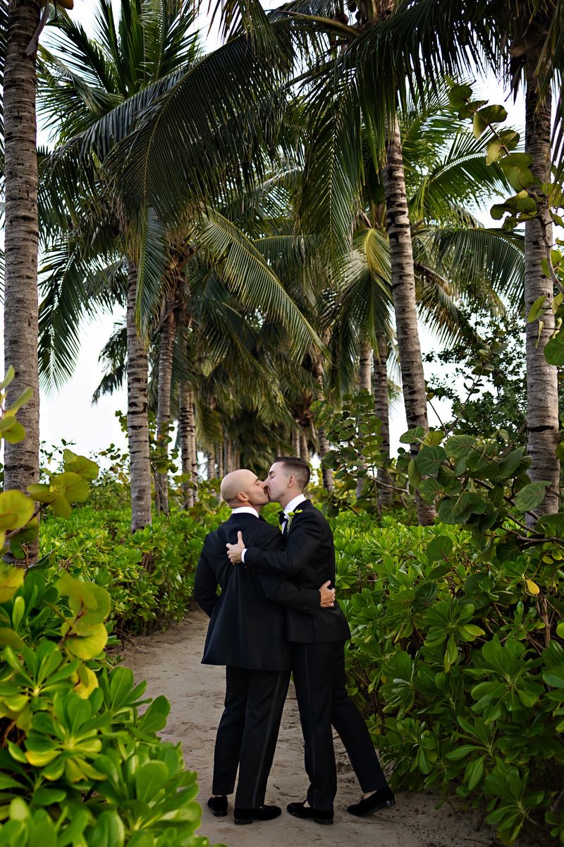 Two grooms share a kiss on a sandy path surrounded by lush tropical palm trees at their outdoor wedding.