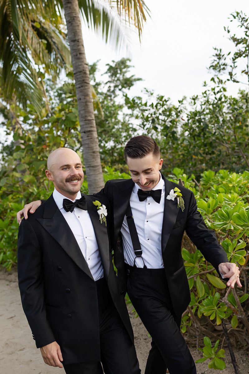 Two grooms in matching black tuxedos with bow ties and boutonnieres smiling together on sandy beach.