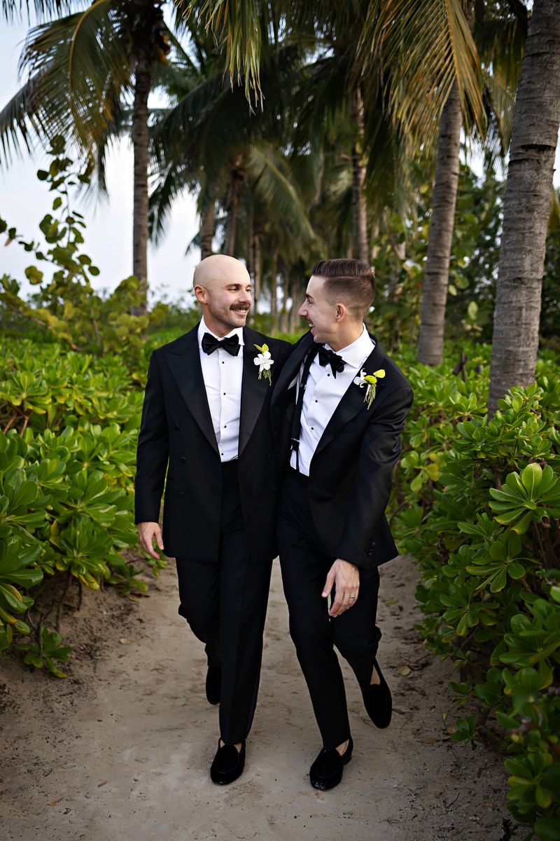 Two grooms in black tuxedos with boutonnieres walk together on a sandy tropical path surrounded by palm trees.