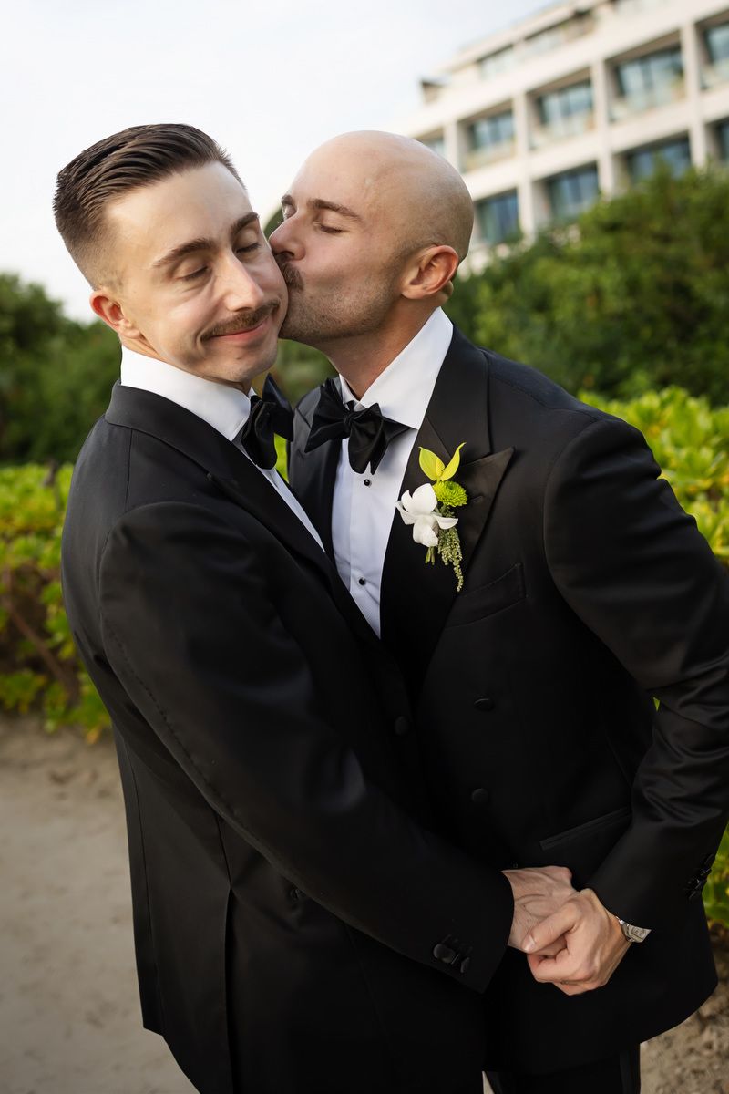 Two grooms in black tuxedos share a tender kiss on their wedding day outdoors.