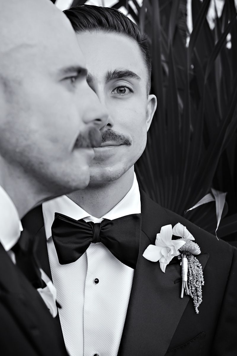 Black and white photo of two grooms in tuxedos with bow ties and floral boutonnieres at a wedding.