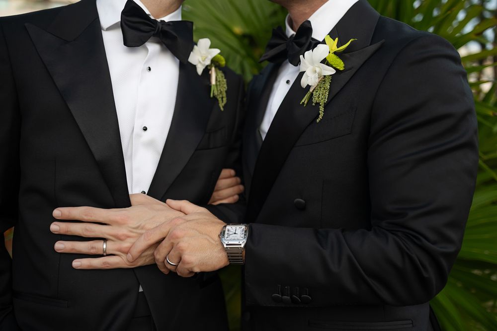 Two grooms in black tuxedos with white floral boutonnieres hold hands showing wedding rings at their ceremony.