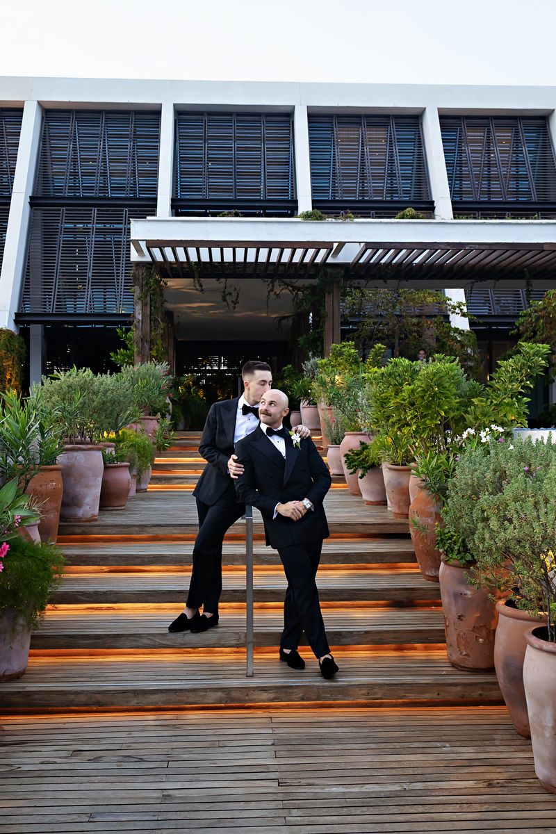Two grooms in black tuxedos embrace on outdoor steps surrounded by potted plants at their wedding venue.