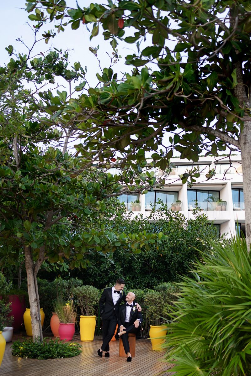 Two grooms in tuxedos share a romantic moment in a lush garden courtyard with colorful planters at their wedding.