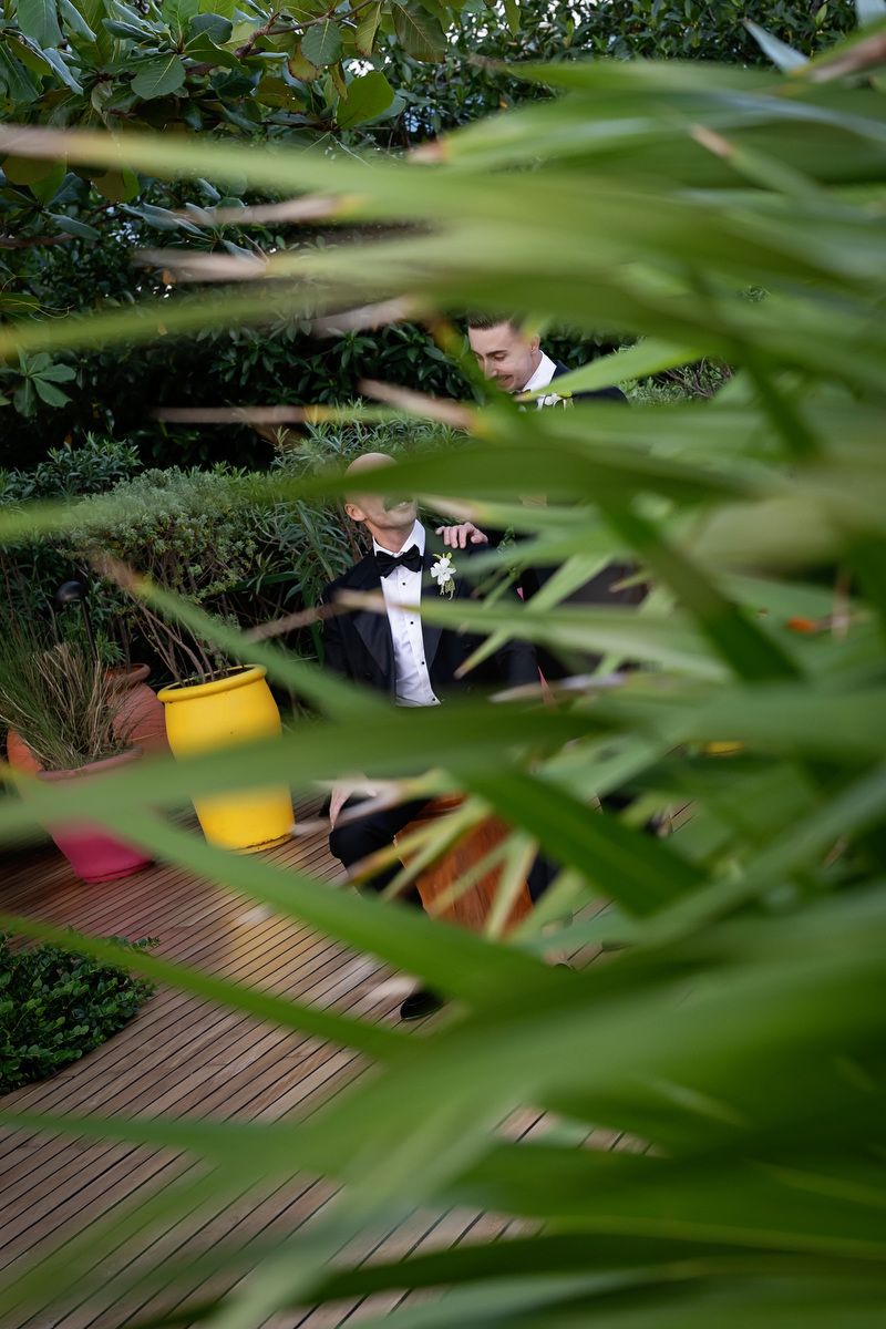 Groom in tuxedo seen through lush green tropical plants on wooden deck at outdoor wedding venue.