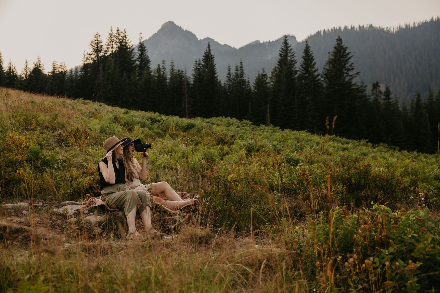 Couple sitting together on a mountain meadow surrounded by evergreen trees and peaks in a scenic outdoor elopement photo.