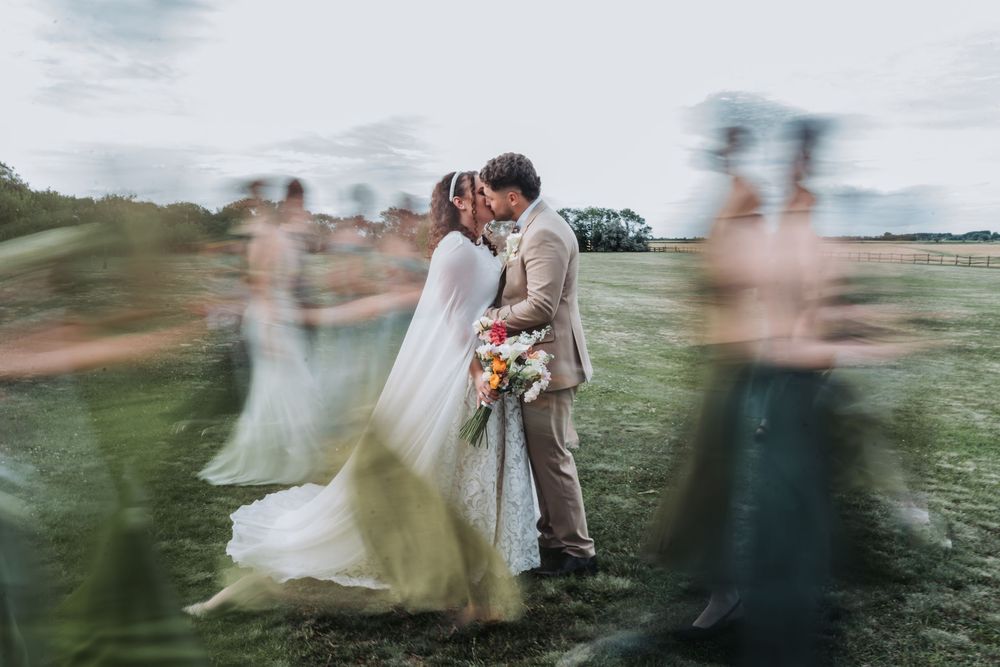 Bride and groom share a kiss outdoors while blurred wedding party moves around them in a dreamy motion effect.