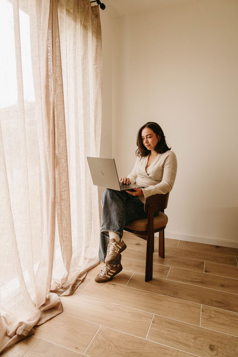 Young woman sitting on wooden chair using laptop by sheer curtain window in bright room.