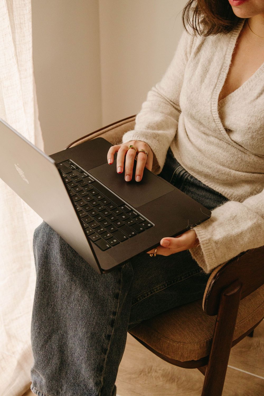 Woman in beige sweater working on laptop while seated in wooden chair near window.