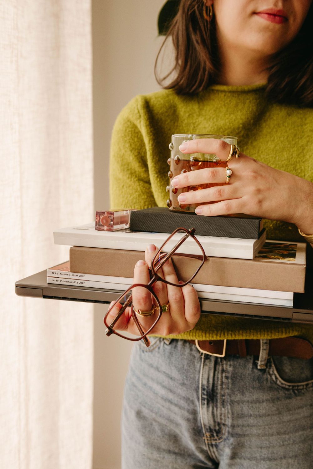 Woman in green sweater holding stack of books, glasses, and glass of tea.