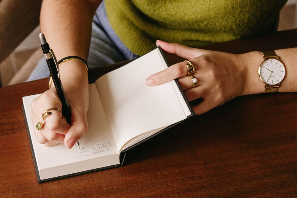 Close-up of woman writing in notebook with pen, wearing gold rings and watch.