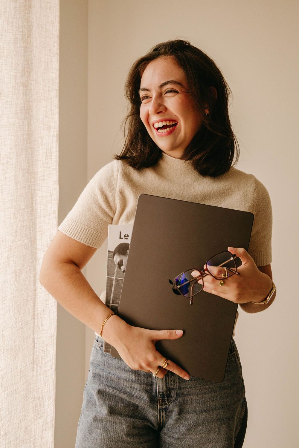 Smiling brunette woman laughing while holding laptop and glasses near window.