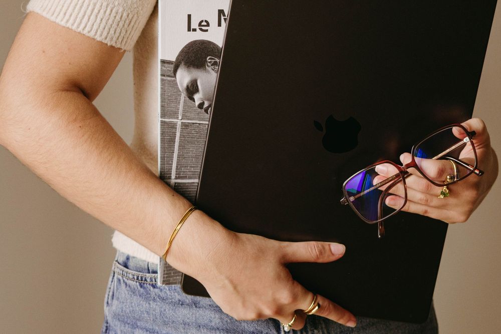 Close-up of person holding black MacBook laptop and glasses with gold bracelet.