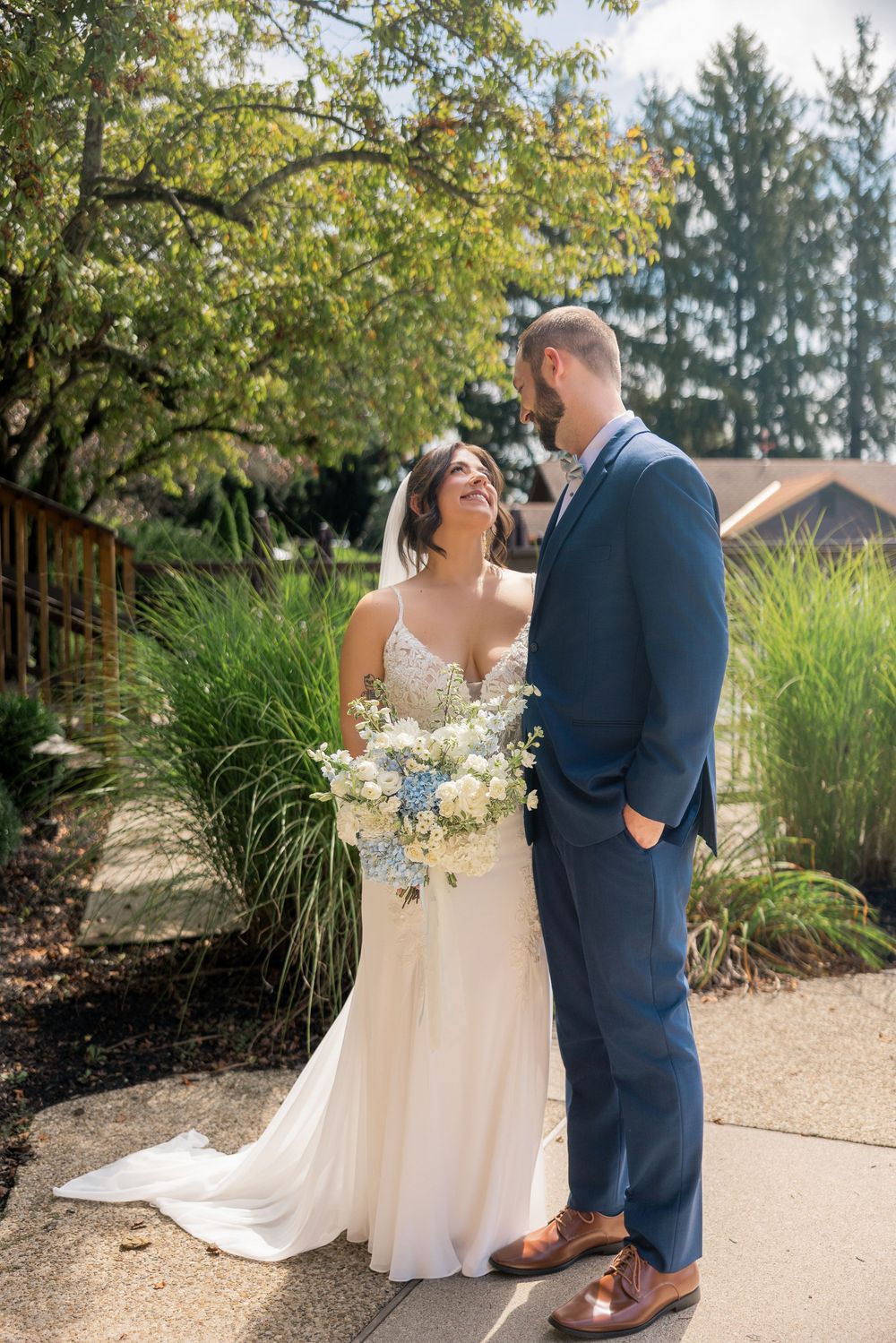 Bride in white gown holding blue and white bouquet smiles at groom in navy suit outdoors on sunny wedding day.