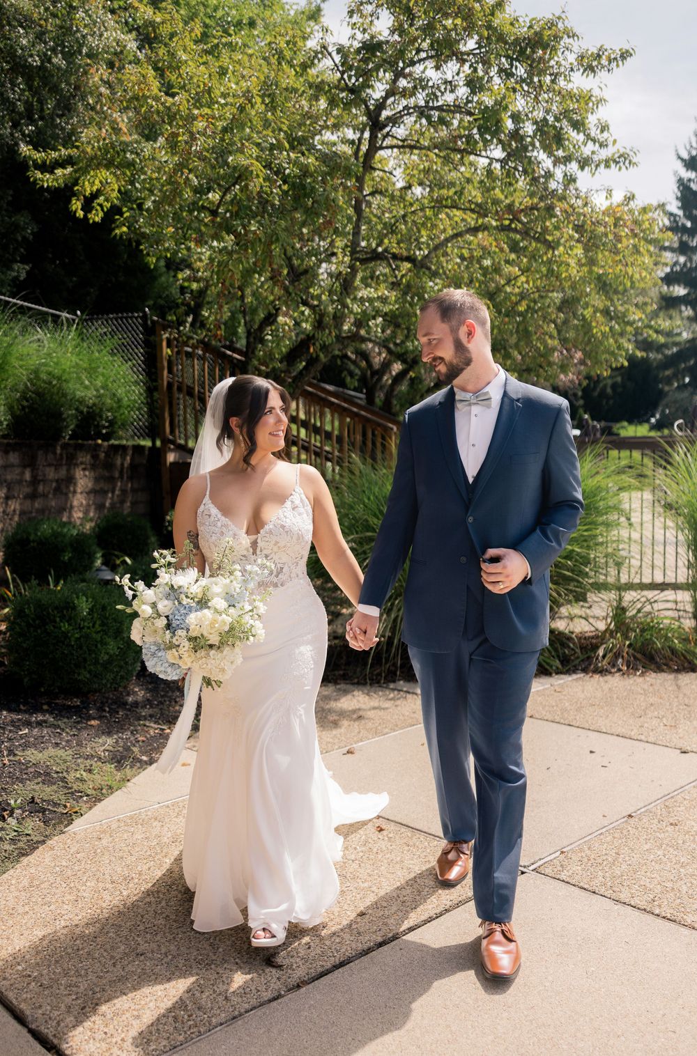 A bride and groom walk hand-in-hand outdoors, she in a white gown with bouquet, he in a navy suit with pink tie.