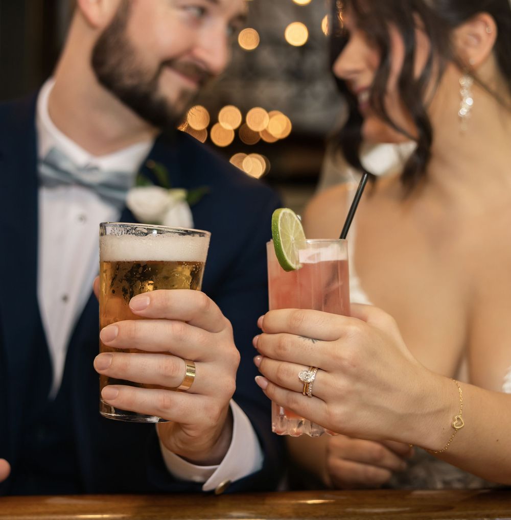 Bride and groom toasting with drinks at wedding reception, groom holding beer, bride holding pink cocktail.