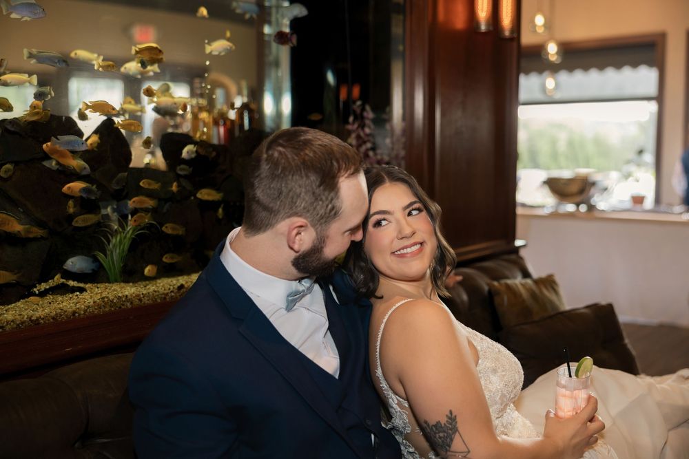 Groom in navy suit kisses bride in lace dress on cheek as she smiles, seated near a fish tank at wedding reception.