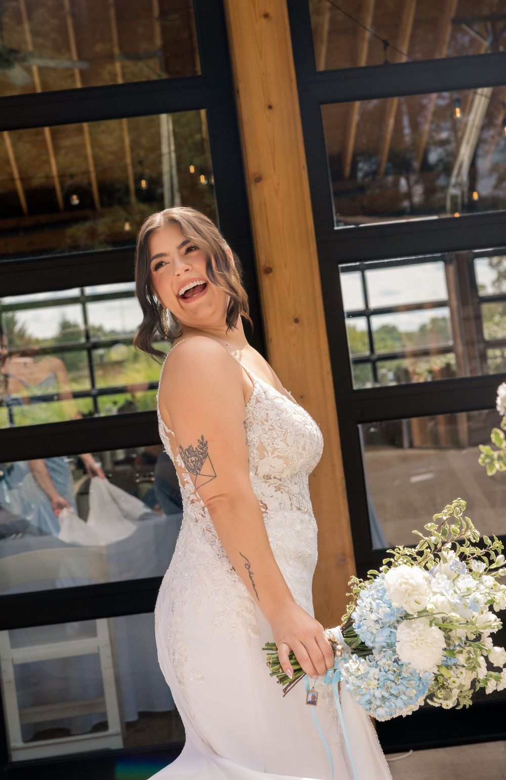 Laughing bride in lace mermaid gown holds blue and white bouquet, standing before large windows at wedding venue.