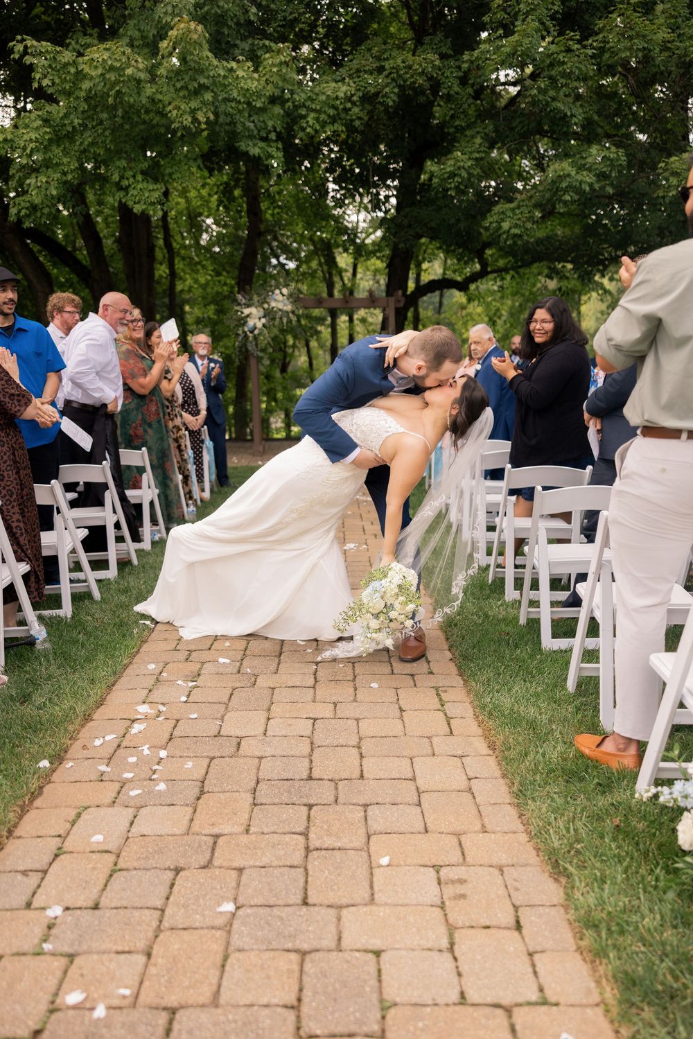 Groom dips bride for a kiss while walking down the outdoor wedding aisle, surrounded by cheering guests seated in white chairs.