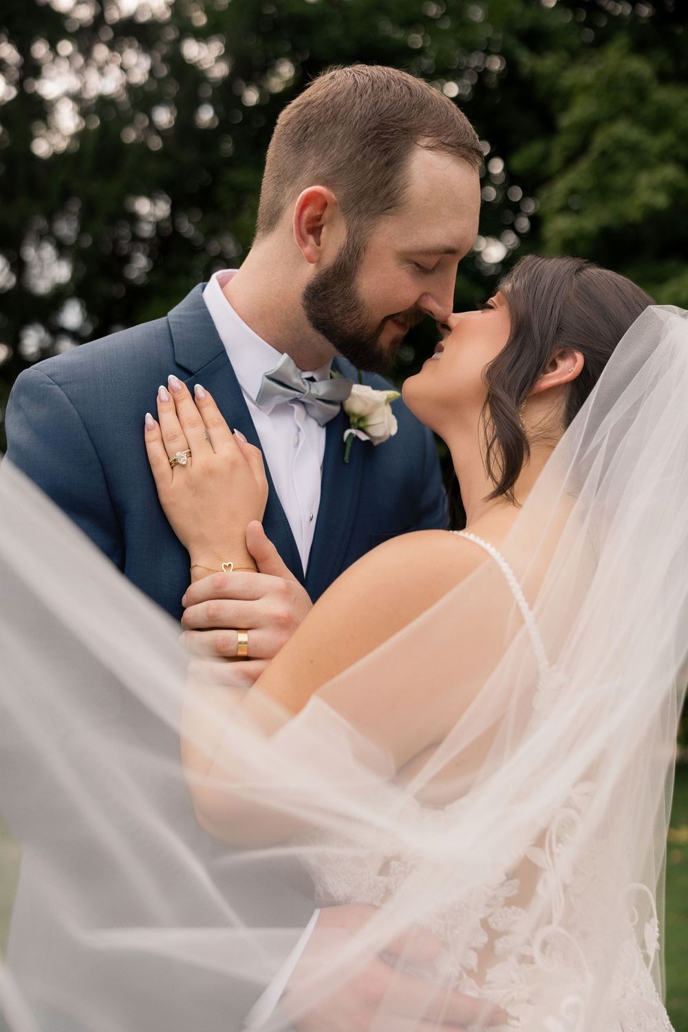 A bride and groom share a romantic almost-kiss, with her veil flowing in the foreground at an outdoor wedding.