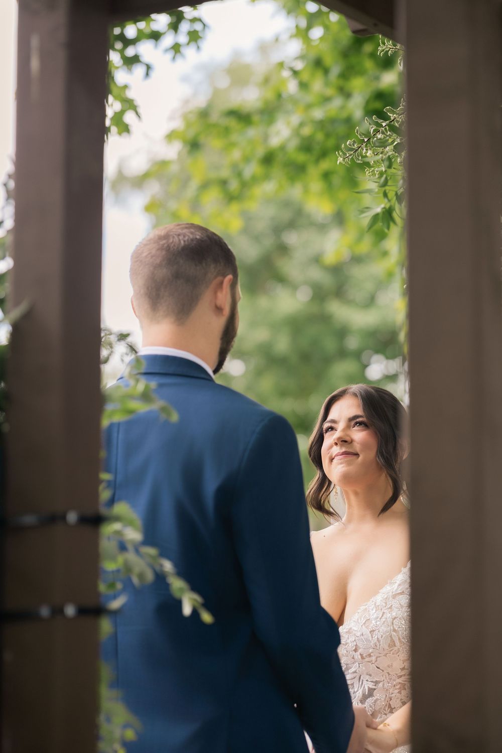 Bride in lace gown smiles at groom in blue suit during outdoor wedding ceremony framed by wooden arch and green trees.