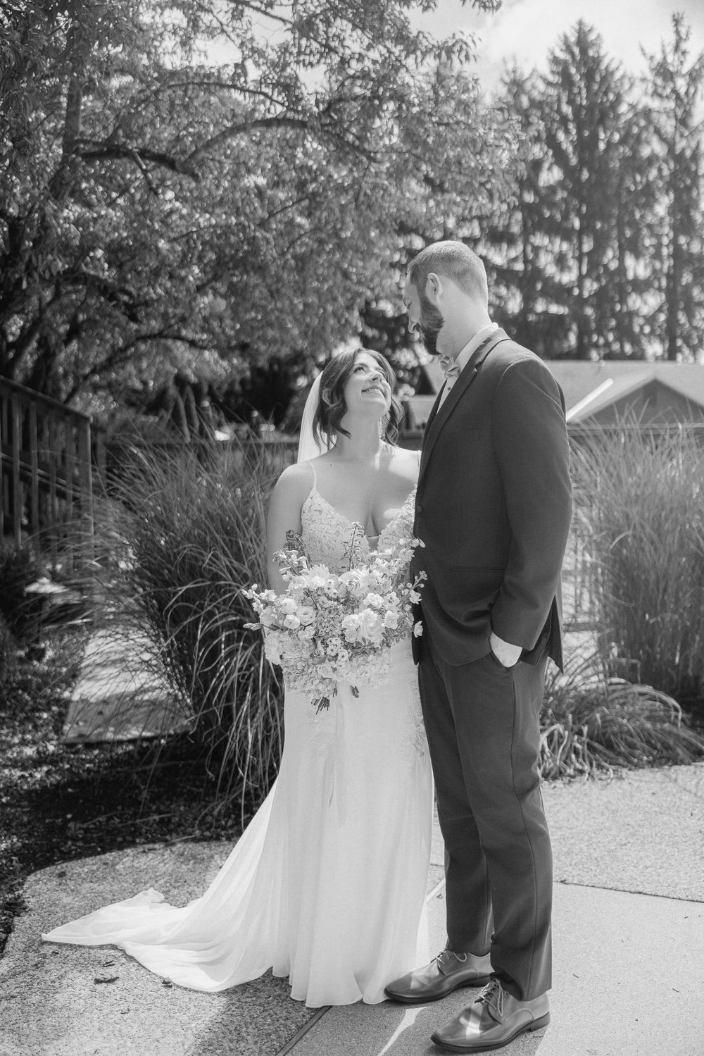 Black and white photo of a bride holding a bouquet and groom in a suit sharing a romantic moment outdoors.