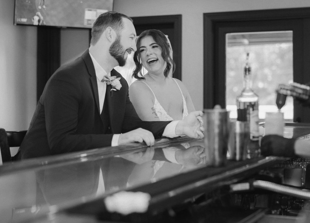 Black and white photo of a laughing bride and groom sharing a joyful moment together at a bar on their wedding day.