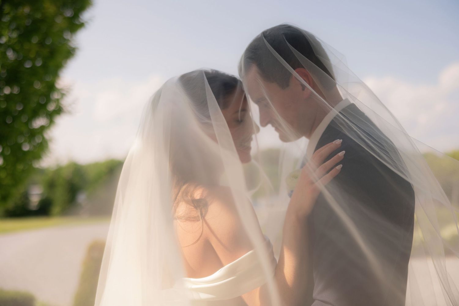 Bride and groom share an intimate moment under a flowing white veil outdoors on their wedding day.