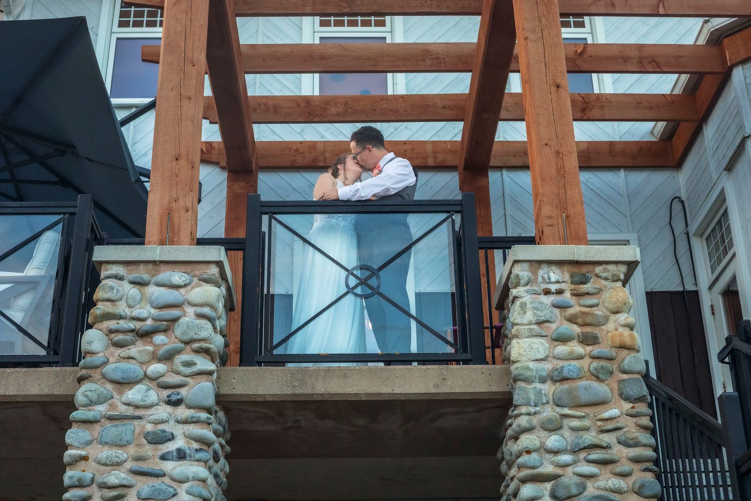 Bride and groom share an intimate moment on a rustic wooden balcony framed by stone columns and exposed timber beams.