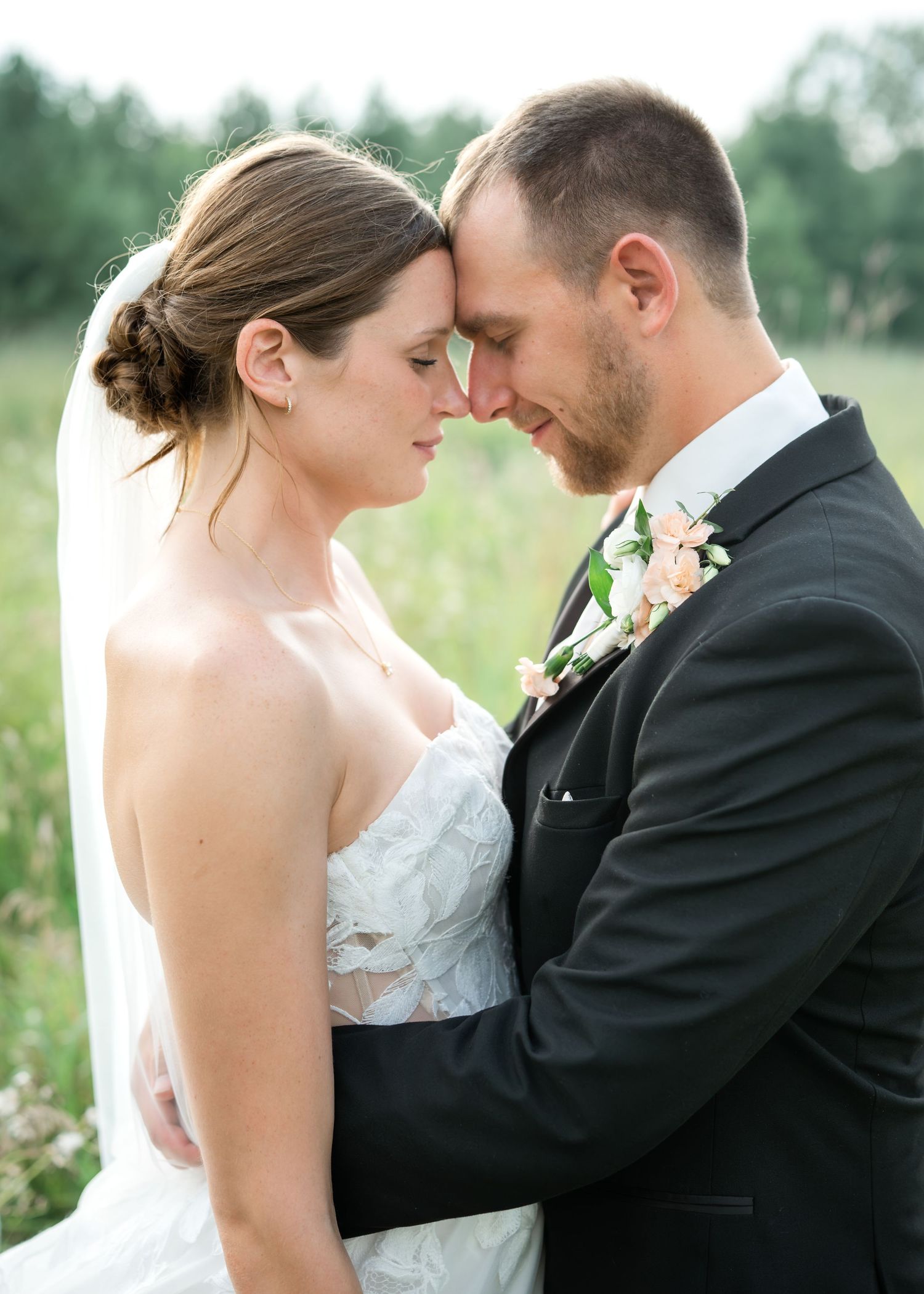 Bride in a white lace gown and veil touches foreheads with her groom in a dark suit and floral boutonniere in a sunlit field.