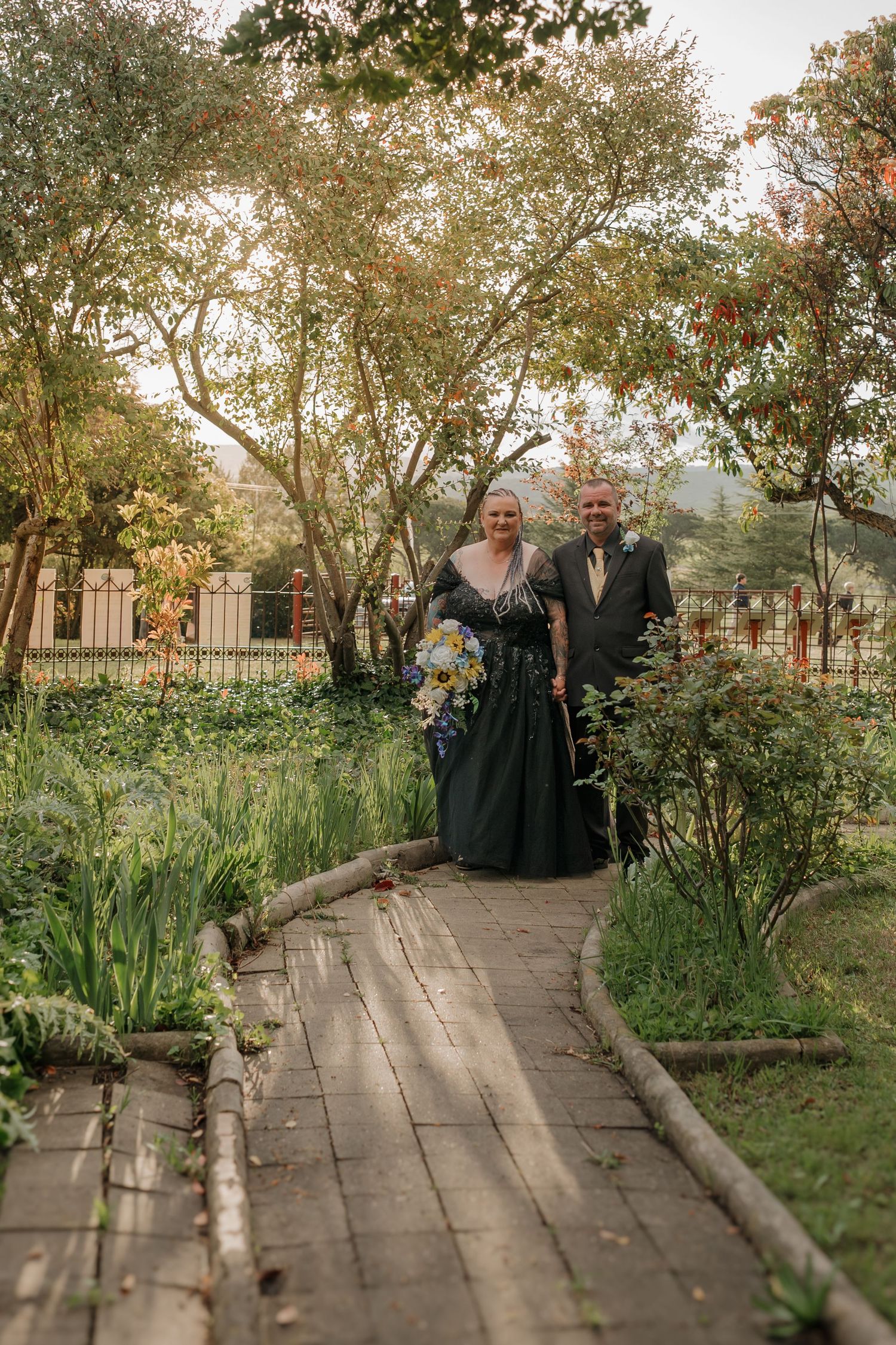 Couple in black formal attire walks garden path, woman holding bouquet, surrounded by lush greenery and trees.